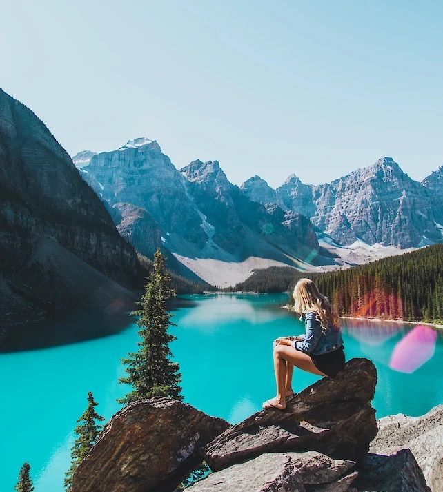 Person sitting on rock overlooking turquoise Moraine Lake surrounded by mountain peaks.