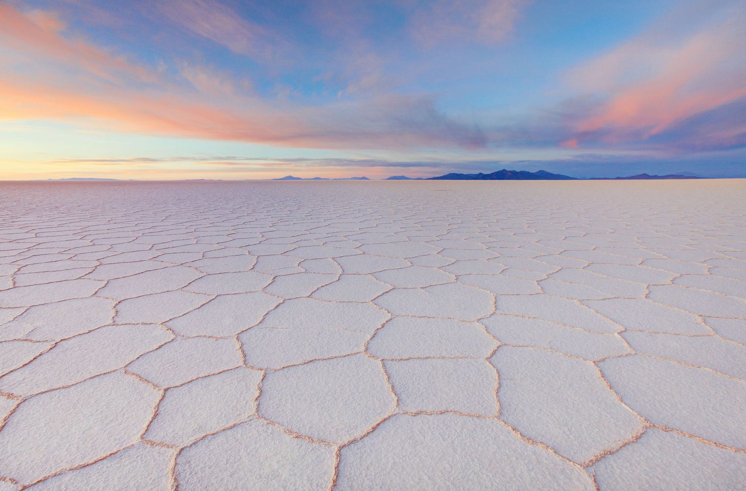 Panoramic view of salt flats with pink and orange sunset sky reflecting on white crystalline surface.