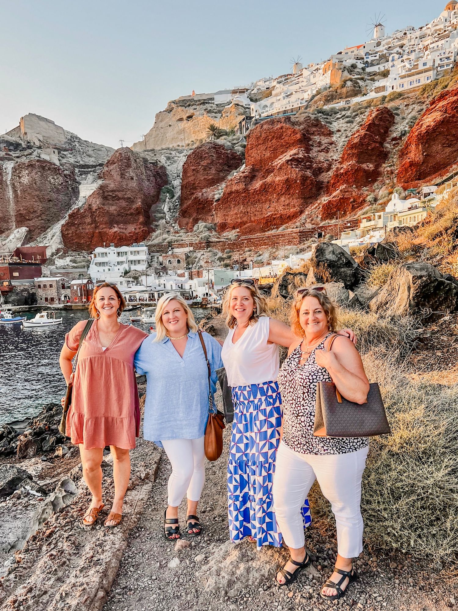 Group gathered on coastal path with Mediterranean village and red cliffs in background at sunset.