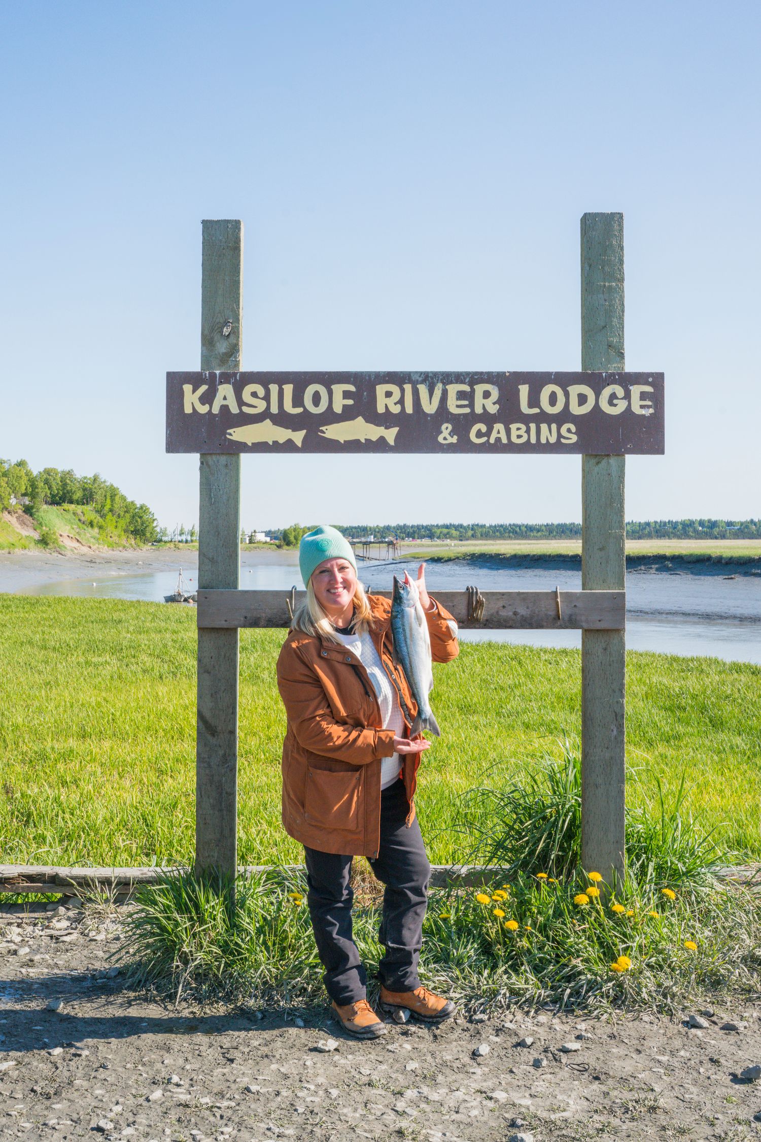 Wooden welcome sign for Kasilof River Lodge & Cabins along a scenic riverbank in Alaska.