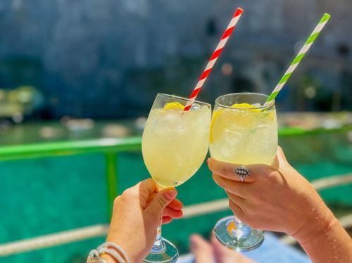 Two people clink glasses filled with a refreshing lemon cocktail garnished with colorful straws on a sunny day.