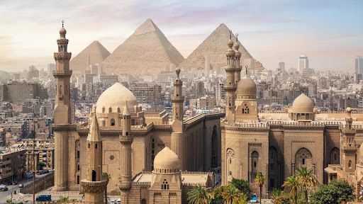 Historic skyline of Cairo featuring ancient mosques, minarets, and pyramids in the distance under a hazy sky.