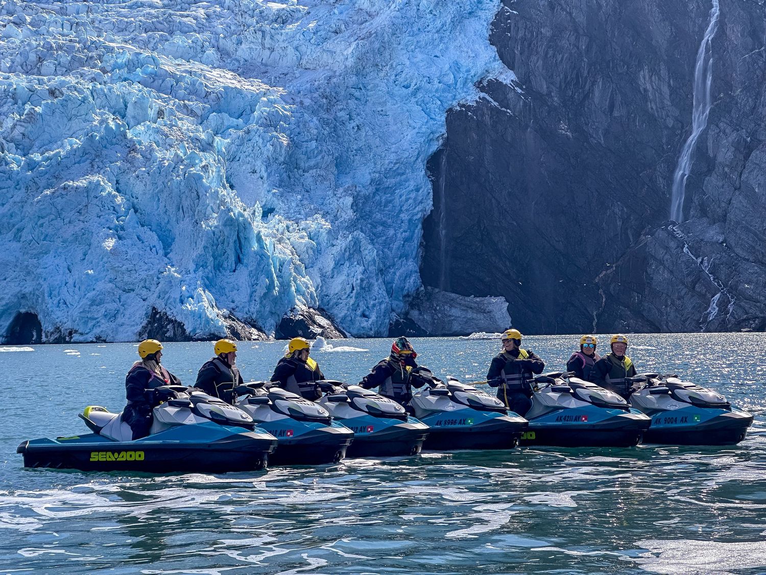 Group of jet skiers lined up in front of a massive blue glacier wall on the water.
