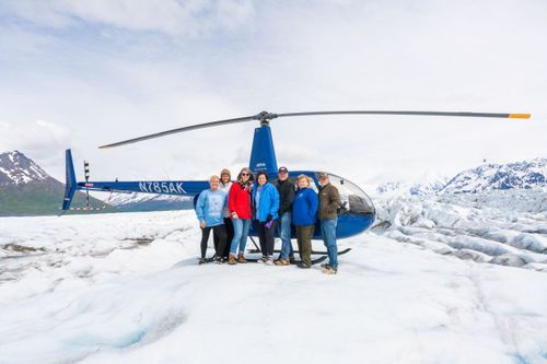Group of adventurers explore a vast glacier landscape with helicopters and snowy mountains in the background.