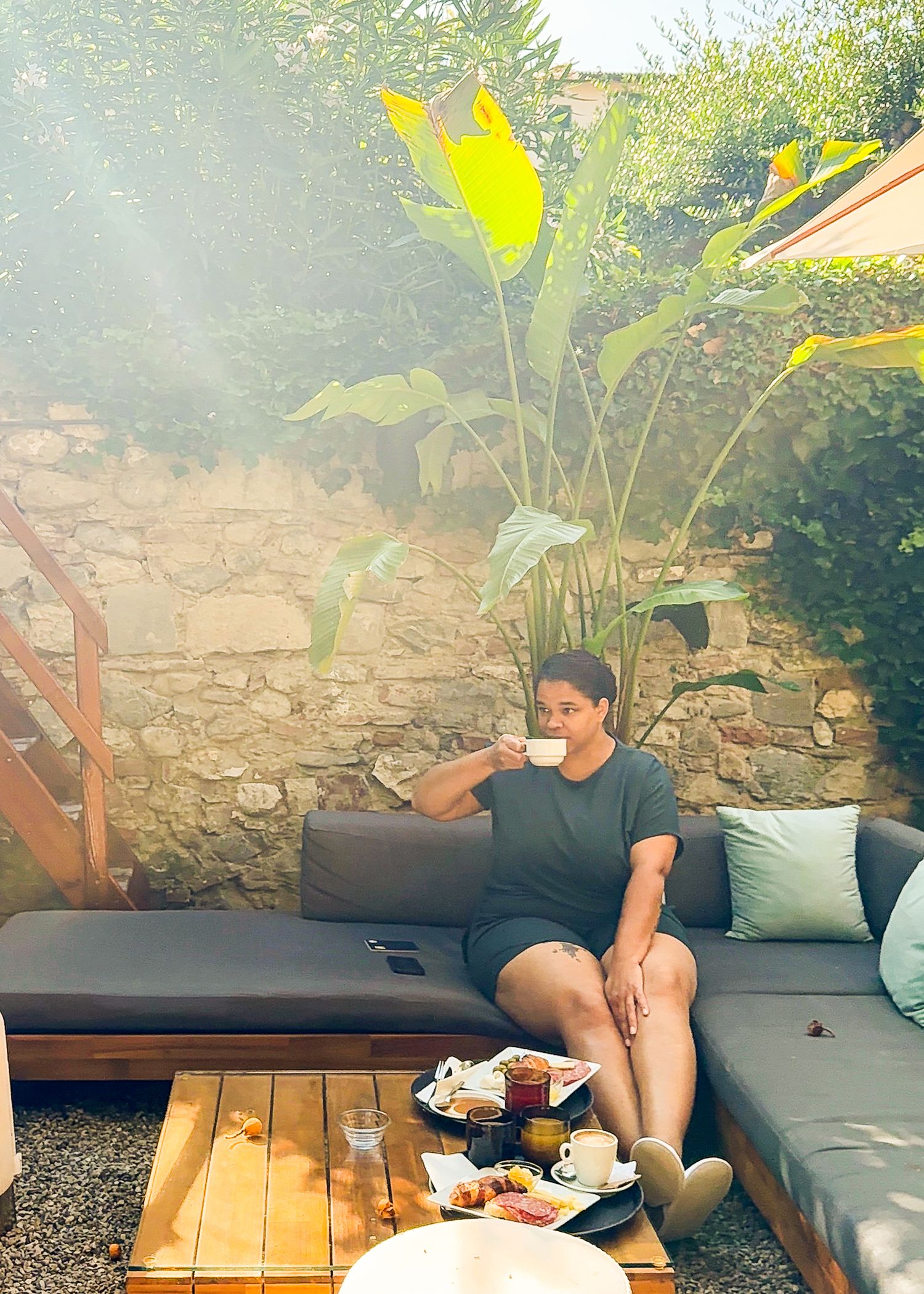 Person relaxing on outdoor patio furniture with snacks under large tropical leaves.