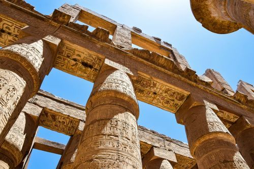 Ancient Egyptian temple columns with intricate hieroglyphic carvings stand against a bright blue sky at Karnak Temple.