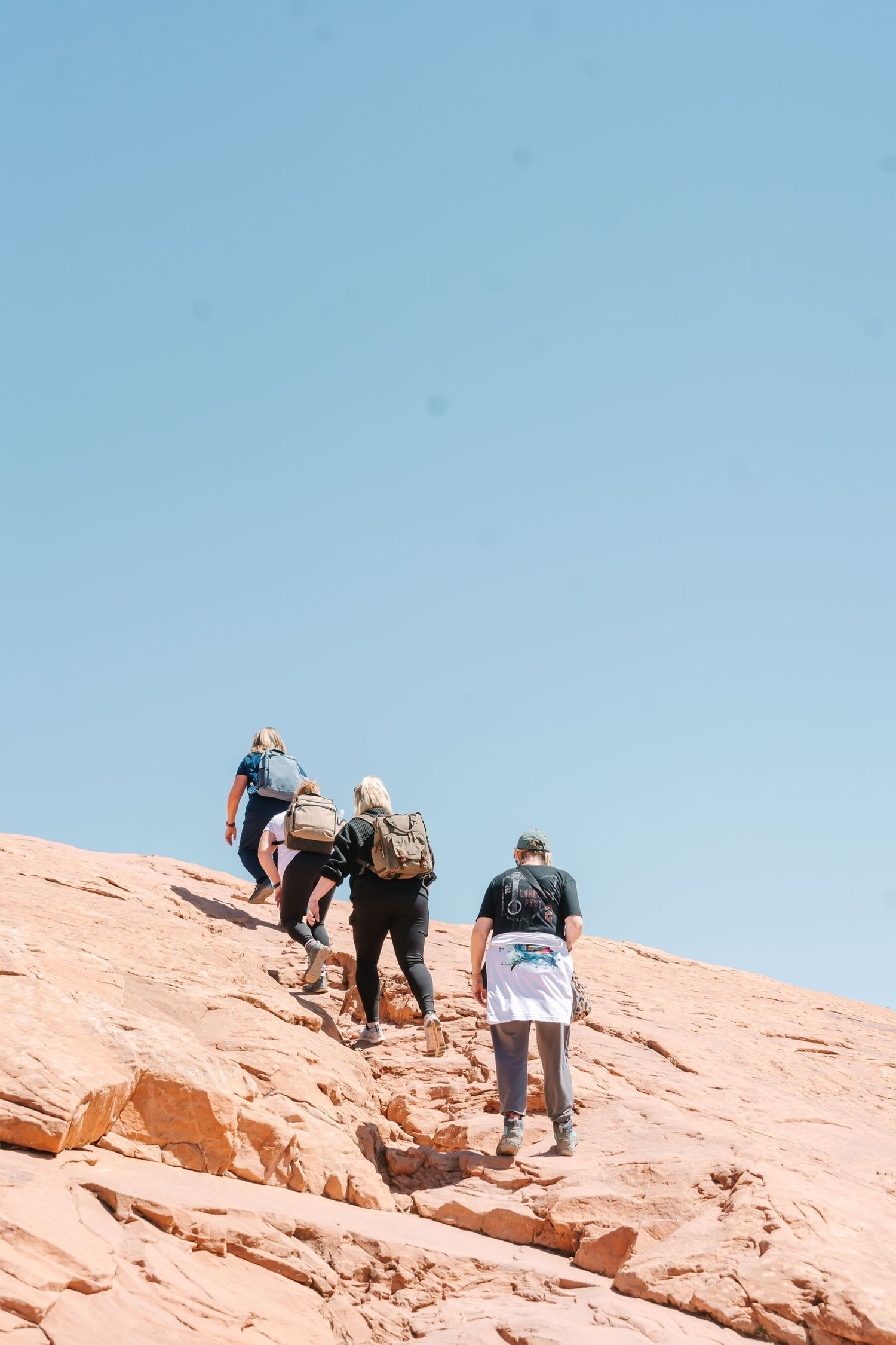 Hikers climbing steep red rock slope under bright blue desert sky.