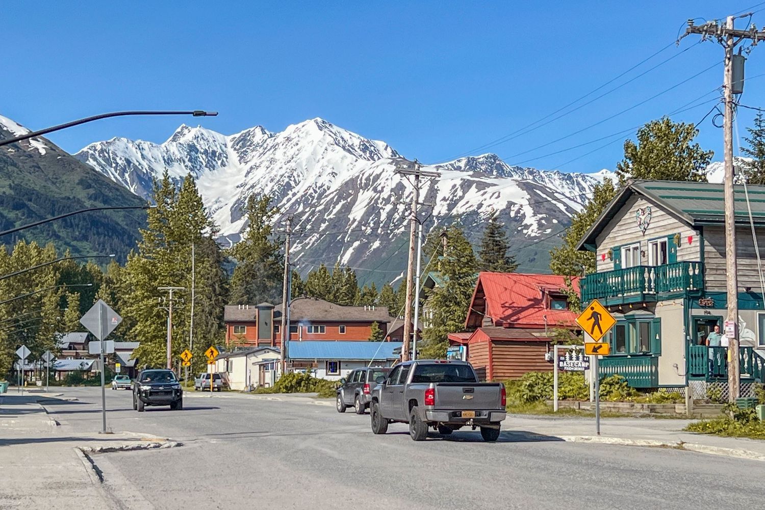 Snow-capped mountains tower over small town buildings and utility poles along a rural street in Alaska.