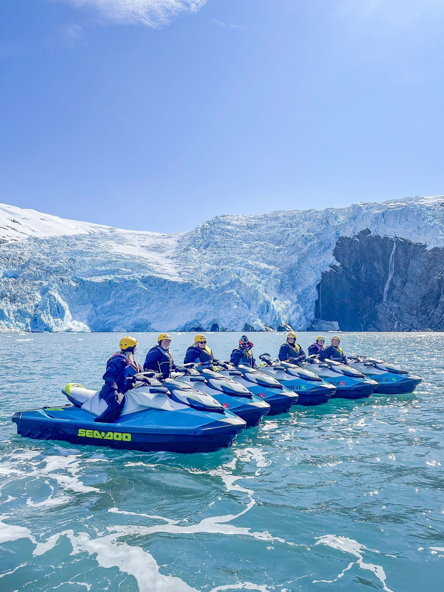 Kayakers in blue vessels navigate through icy waters near a massive glacier under bright blue skies.