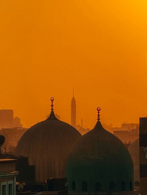 Silhouettes of mosque domes and minarets against an orange sunset sky in Cairo with the iconic tower in background.