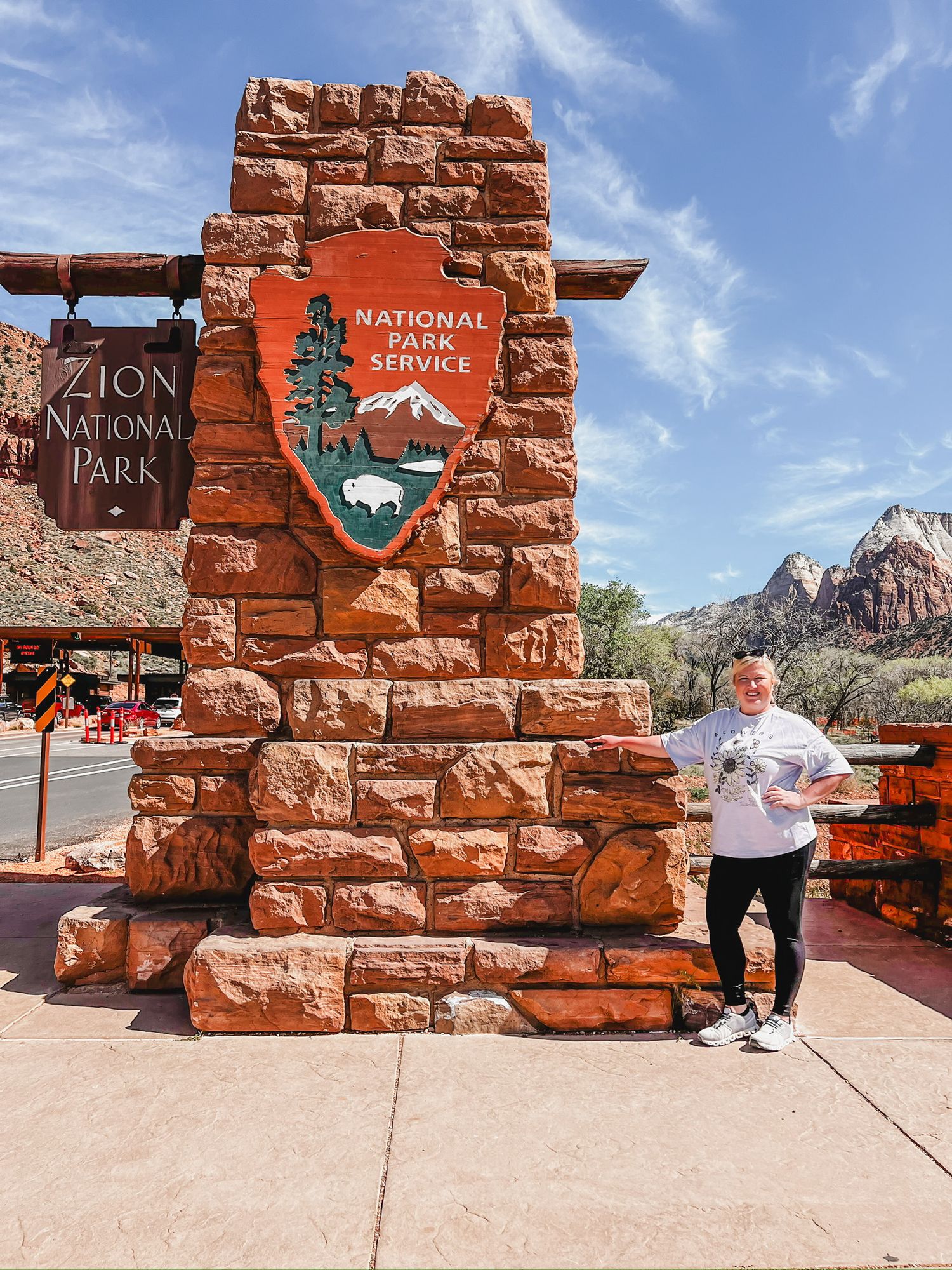 Red sandstone entrance sign for Zion National Park with desert mountains in the background under blue sky.