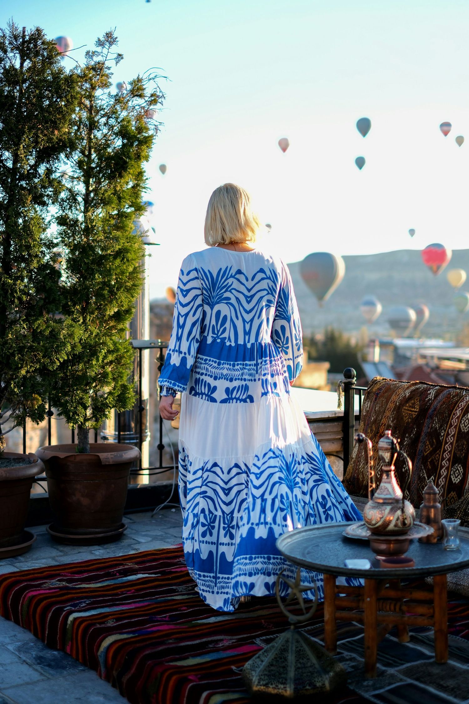 Hot air balloons float in the distance as a figure in blue and white dress enjoys the scenic morning view.