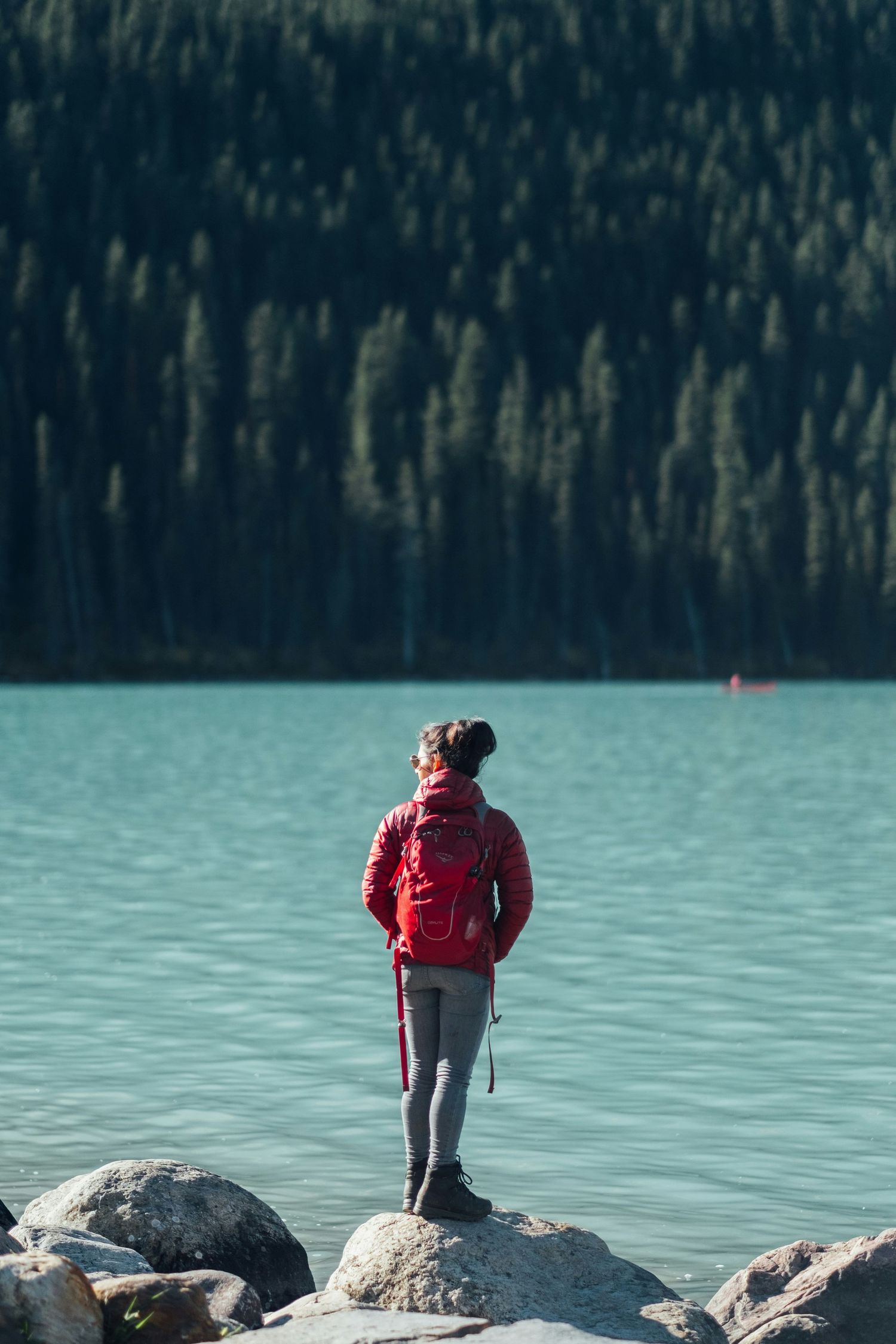 A person in a red jacket stands on large rocks along the turquoise waters of a serene mountain lake.