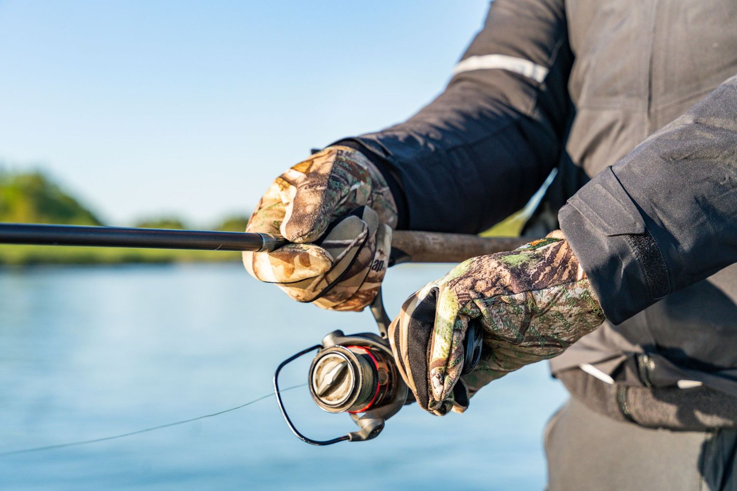Close-up of fishing hands holding rod with camouflage gloves on calm water.