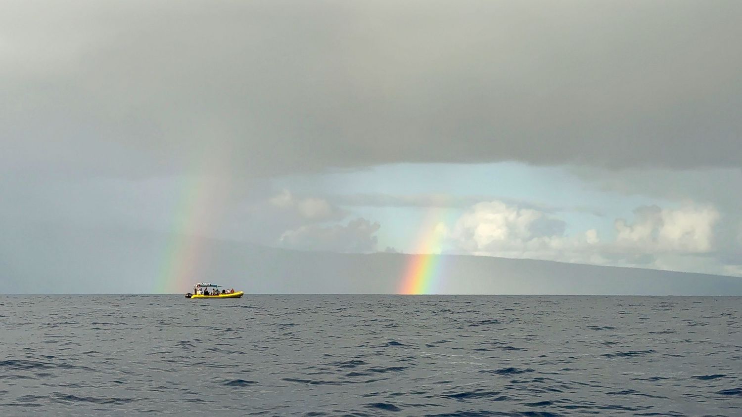 Scenic ocean horizon at sunset with a distant boat and rainbow visible over the water.