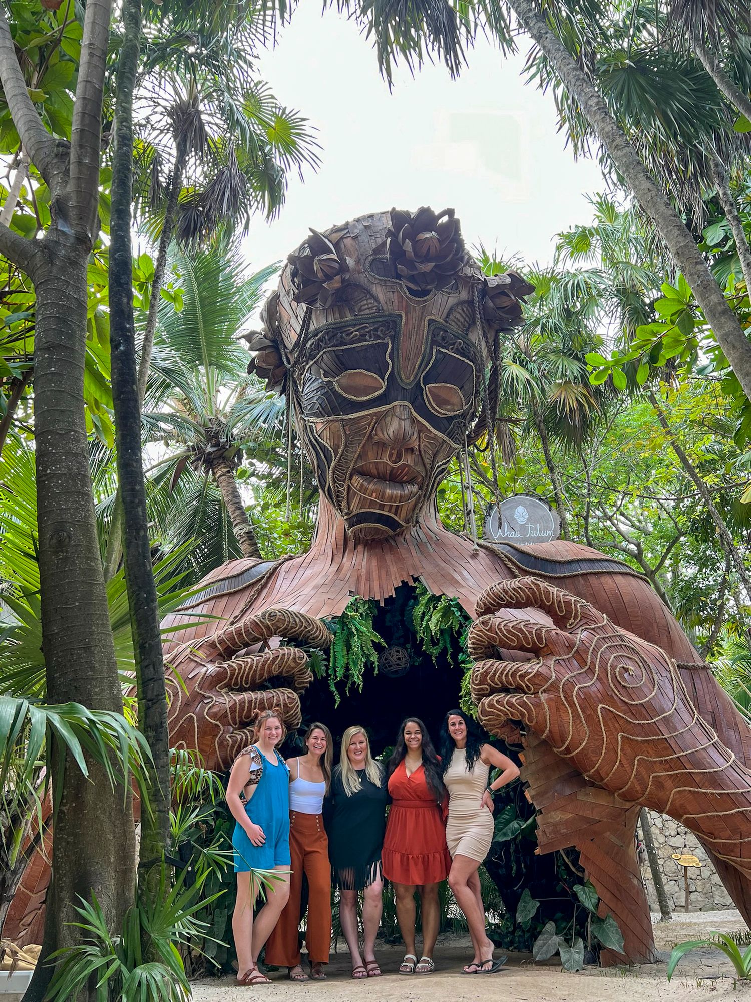 Carved wooden troll statue installation among tropical palm trees in a garden setting, Tulum, Mexico.
