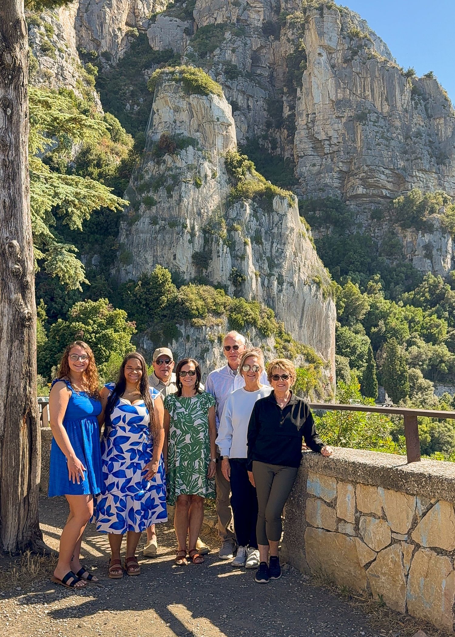 Group of tourists enjoying scenic mountain views from a stone terrace overlook in a Mediterranean landscape.