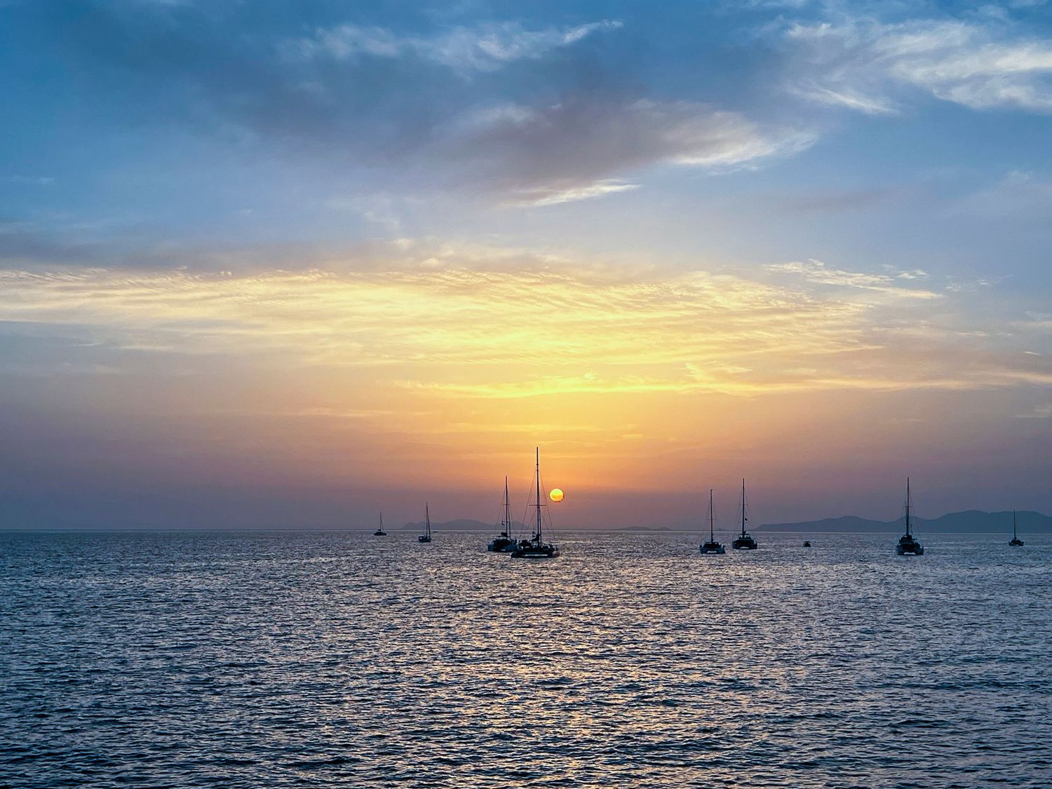 Silhouettes of sailboats dot the horizon during a vibrant orange ocean sunset.