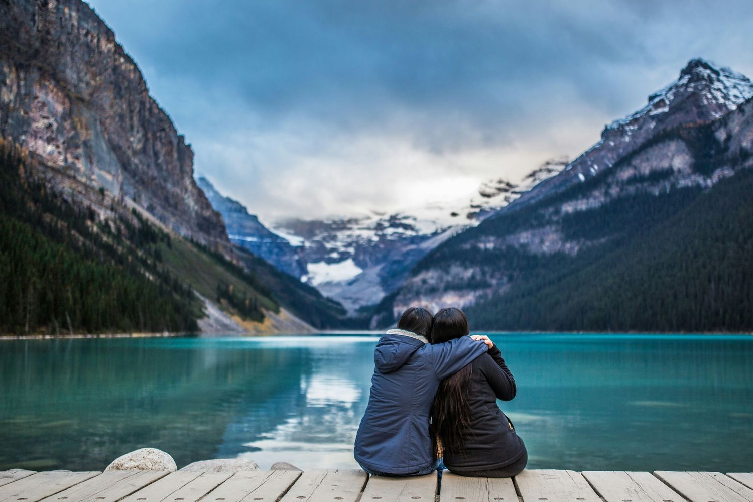 Two people sit together on a wooden dock overlooking turquoise Lake Louise and snow-capped mountains in Banff National Park.