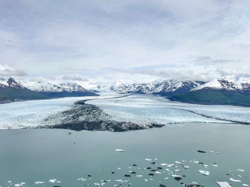A panoramic view of snow-capped mountains and glaciers reflecting in the calm waters of an Alaskan fjord.
