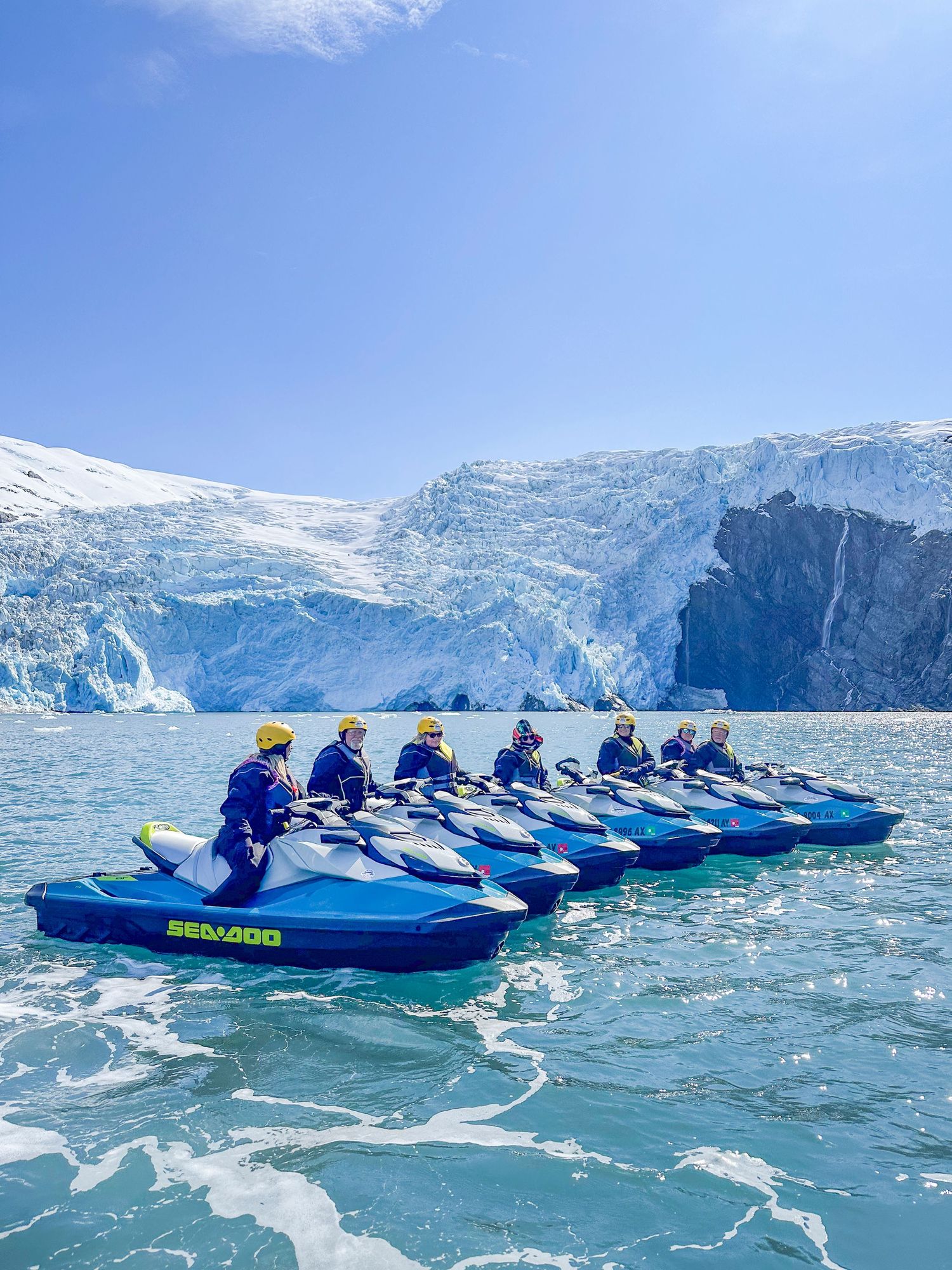 Group of jet-skiers lined up in blue boats near a massive glacier wall.