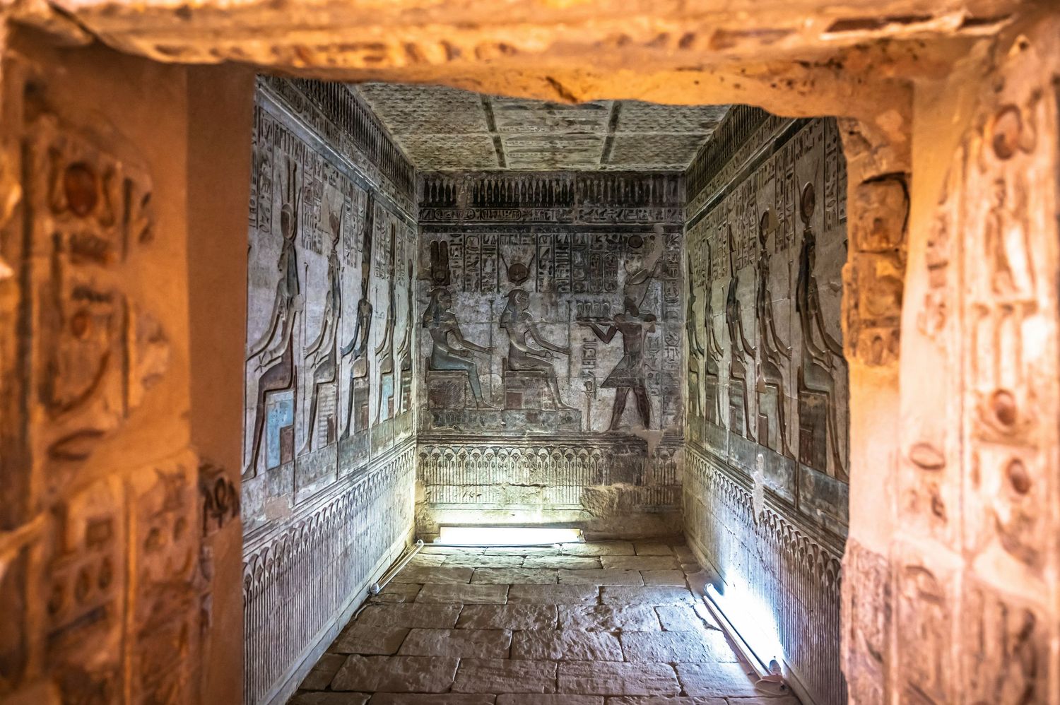 Interior corridor of an ancient Egyptian temple with intricate hieroglyphs and carvings illuminated by natural light and shadows.