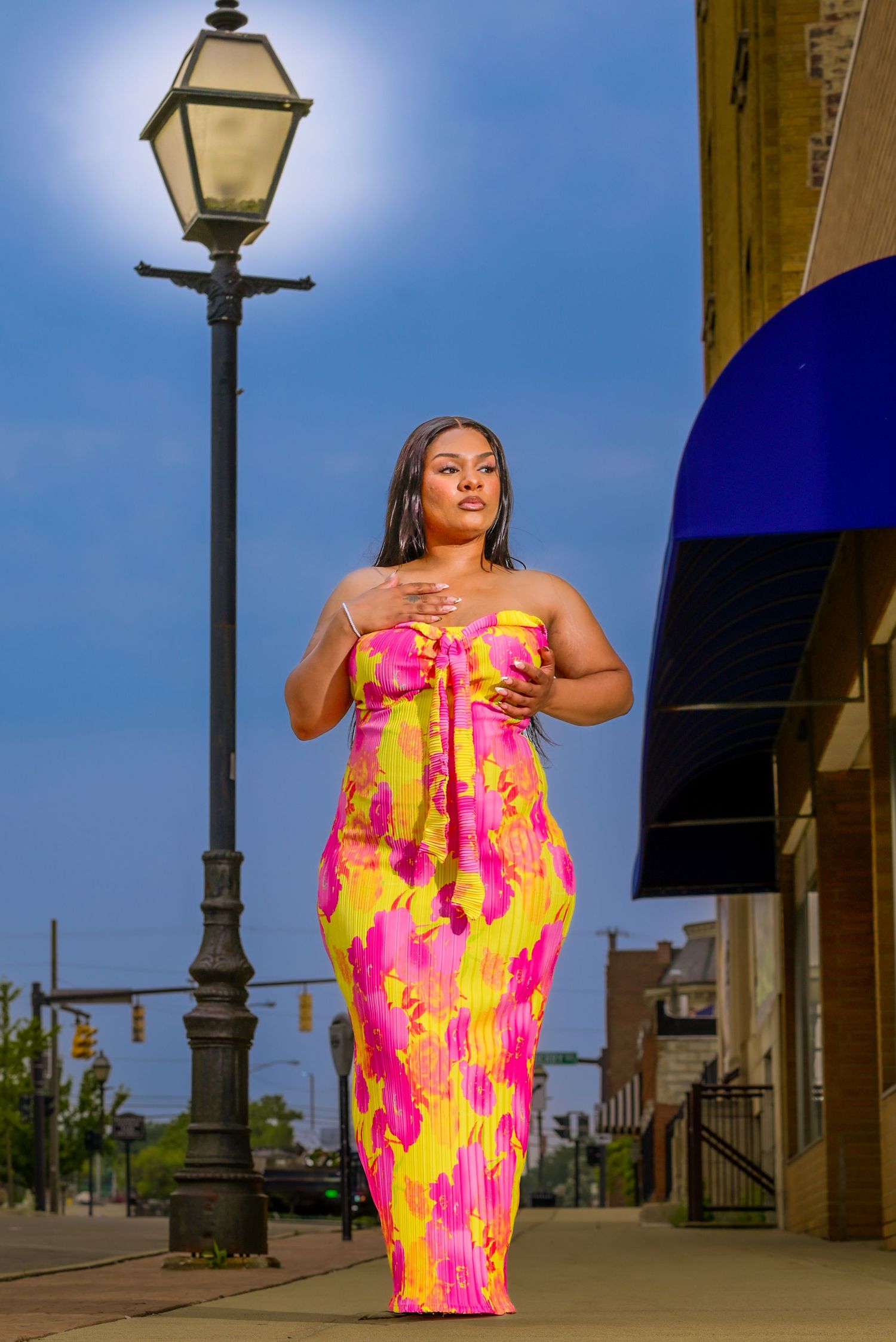 A vibrant pink and yellow tropical print maxi dress worn on a sidewalk with a vintage street lamp and blue awning nearby.