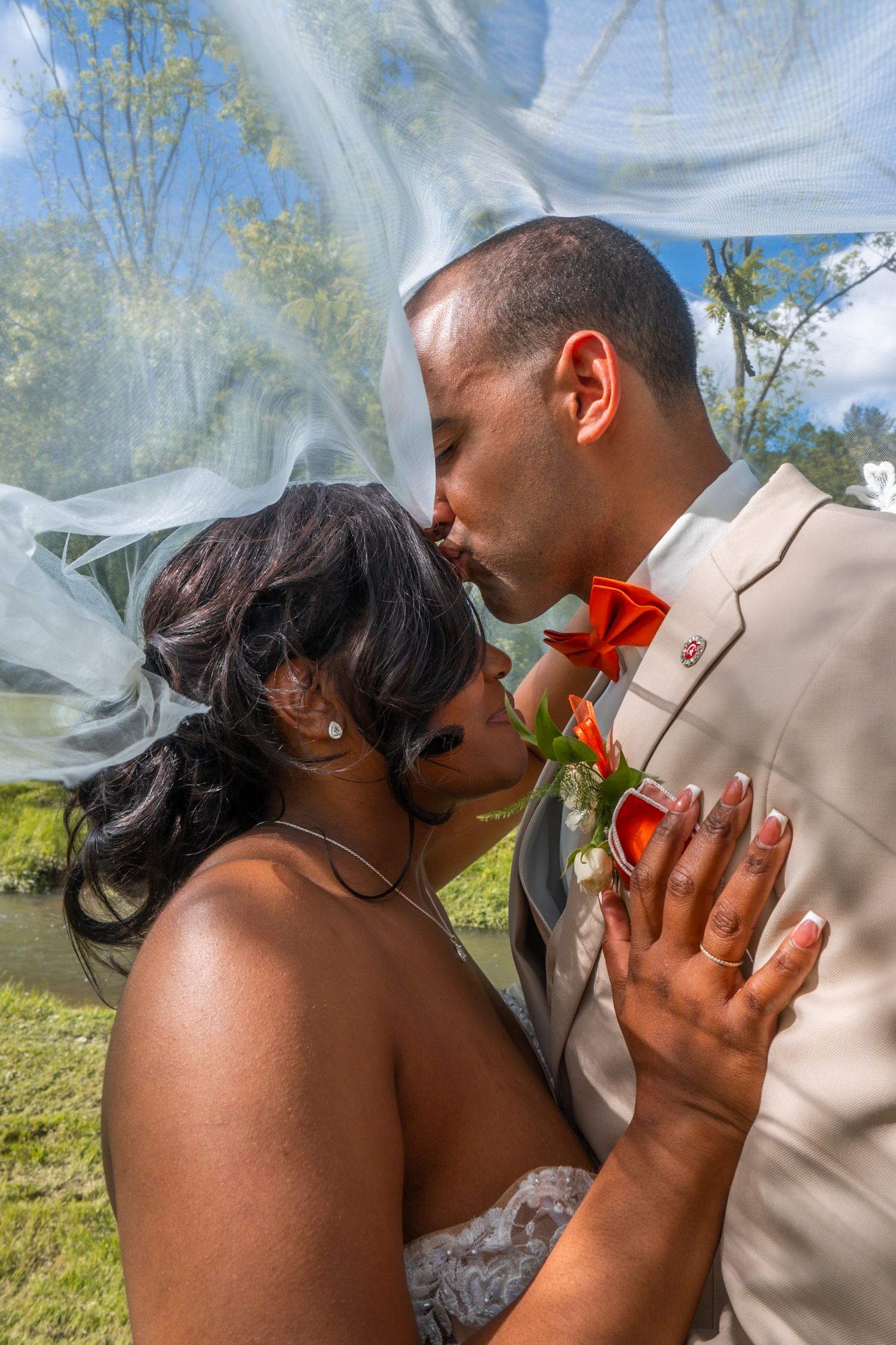 A bride and groom share a romantic kiss outdoors, with a flowing veil and red roses, dressed in wedding attire.