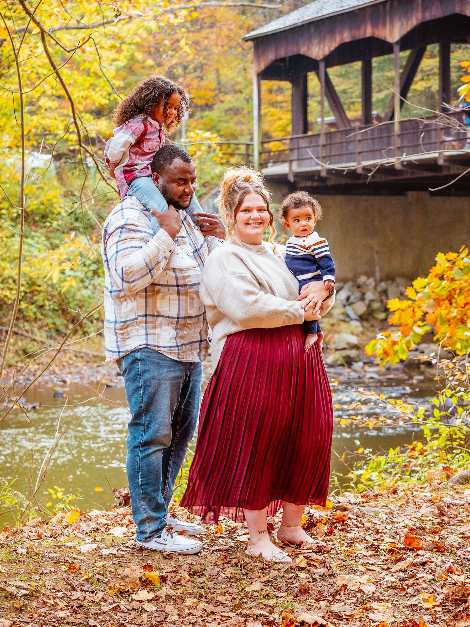 Family of four poses by a covered bridge amid fall foliage, parents holding two young children on an autumn day.