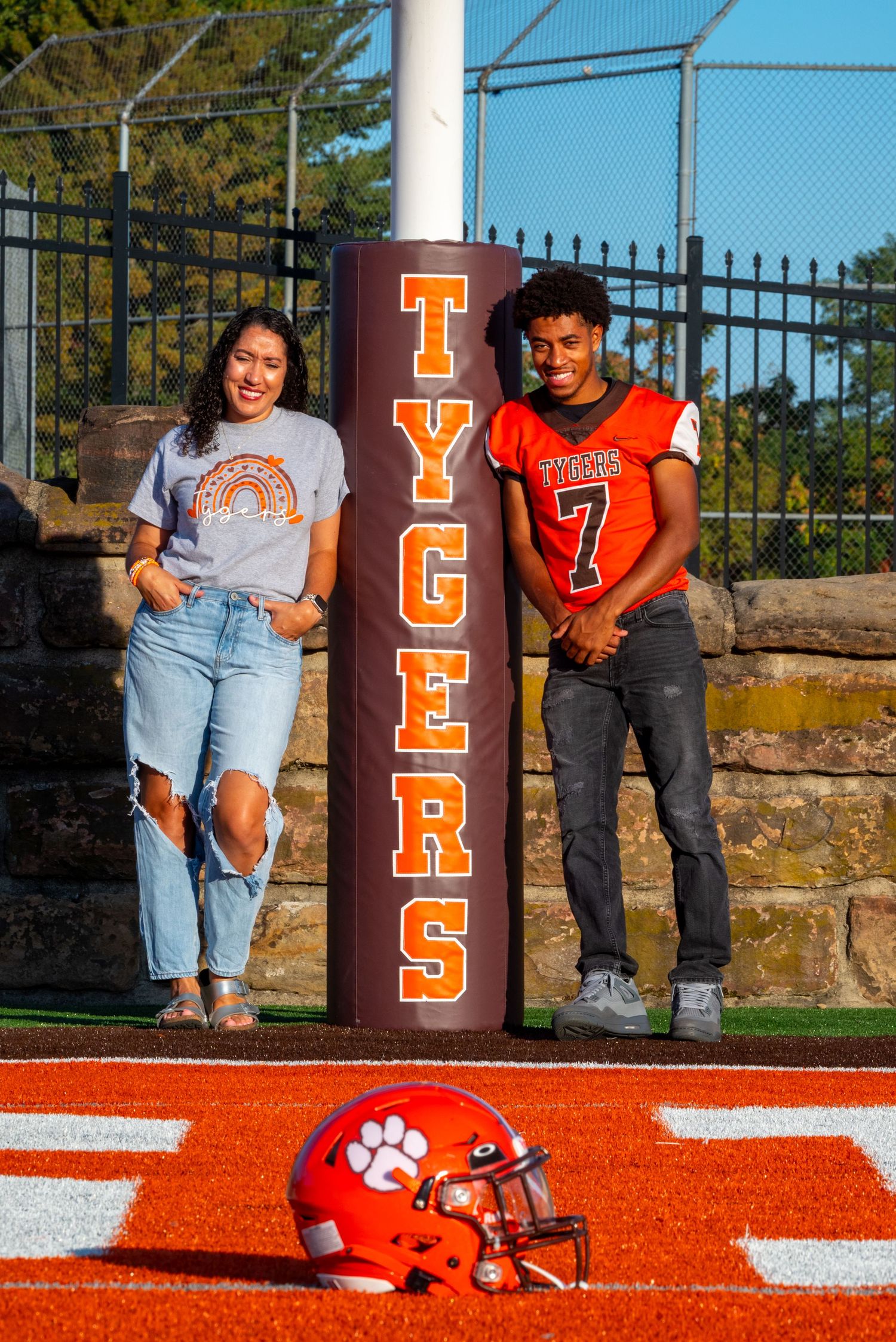 Two students pose beside a Tygers football field goal post with an orange helmet on the turf.
