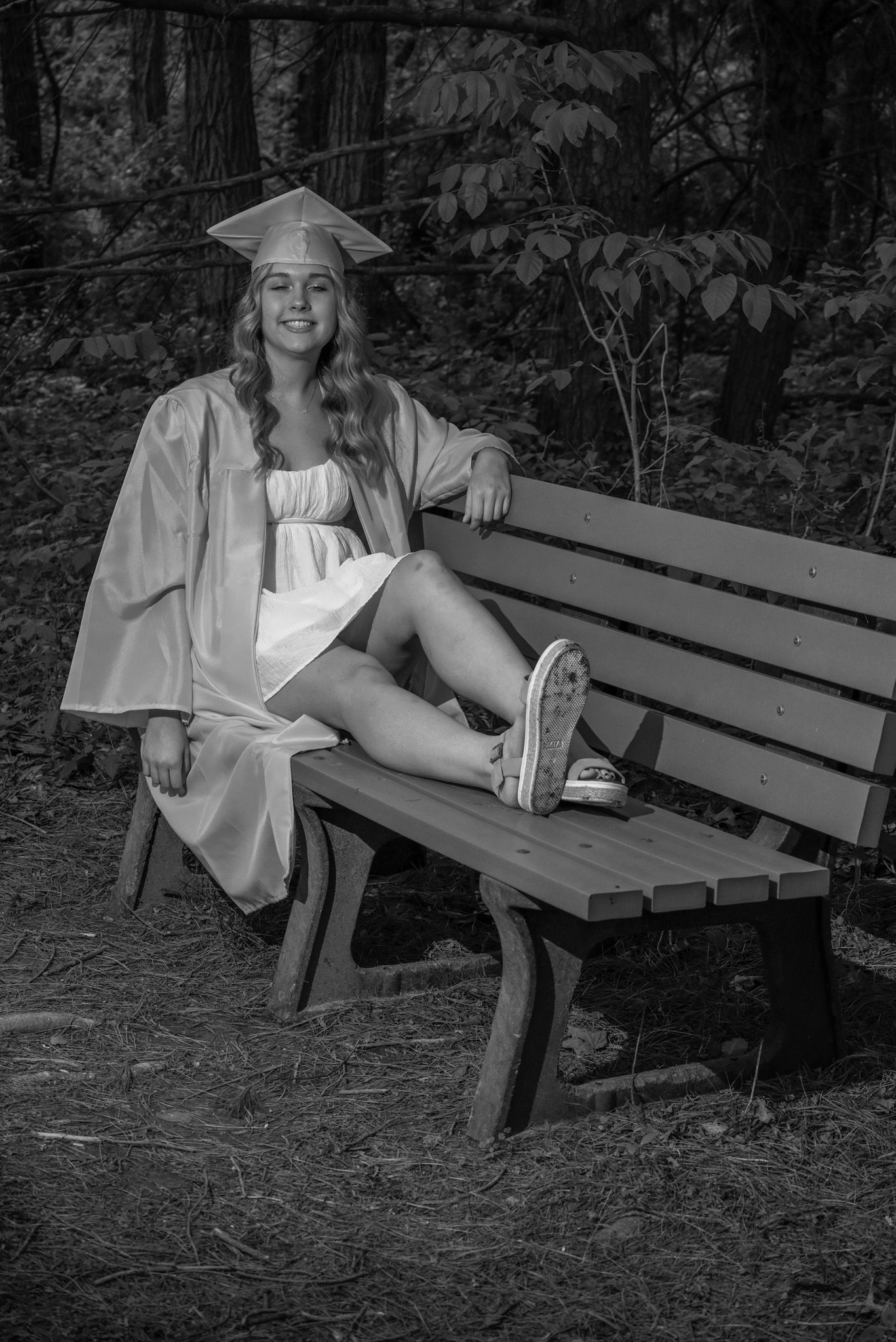 Black and white photo of a smiling graduate in cap and gown sitting on a wooden bench outdoors.