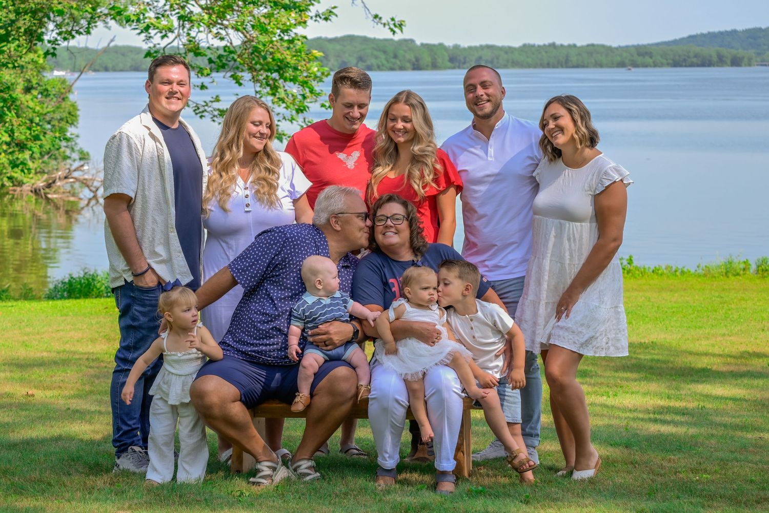 Large family group portrait outdoors by a lake, featuring adults and babies posing together on green grass.