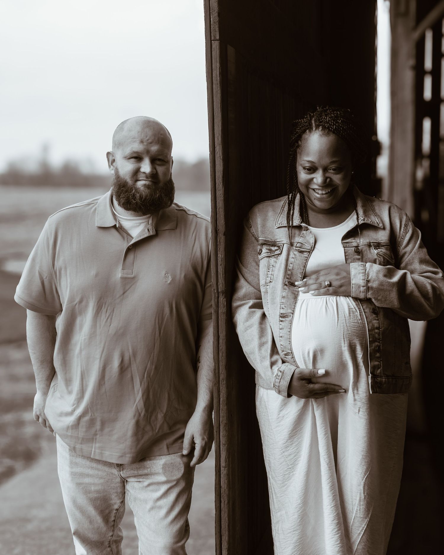 Black and white photo of a smiling pregnant woman in denim jacket and a bearded man standing in a doorway.