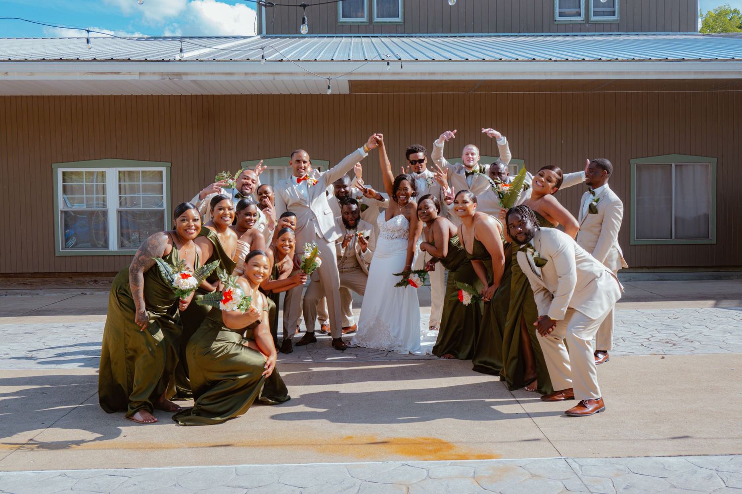 Wedding party celebrating outdoors, bridesmaids in olive gowns and groomsmen in tan suits cheering behind bride and groom.