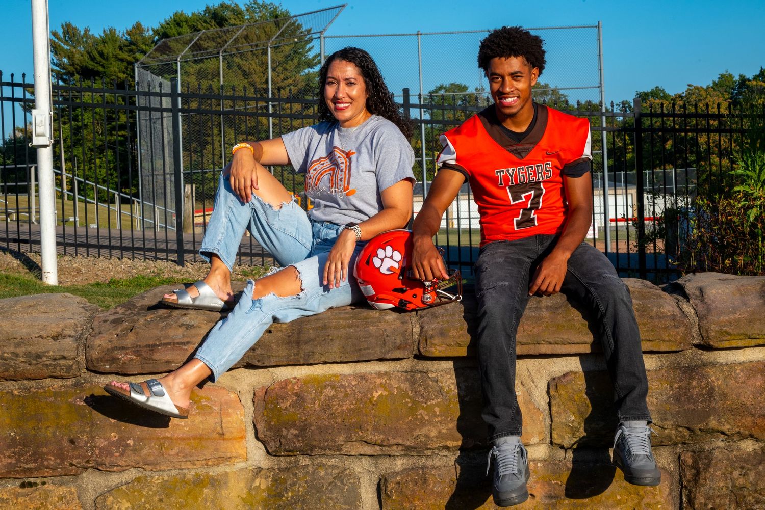 Two high school students in sports attire sit on a stone wall near a football field on a sunny day.