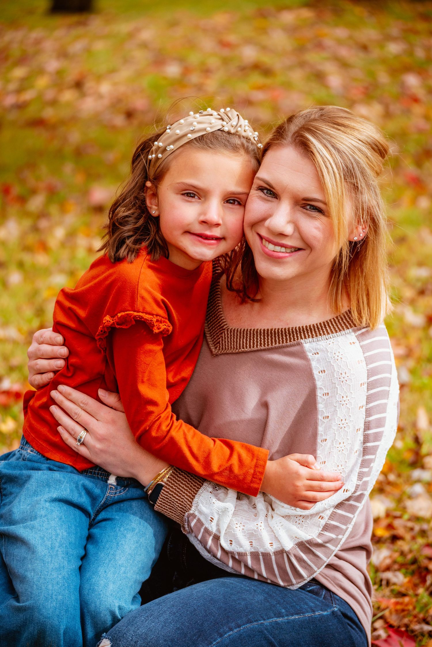 A woman and young girl hugging outdoors in autumn, surrounded by colorful fall leaves in a park setting.