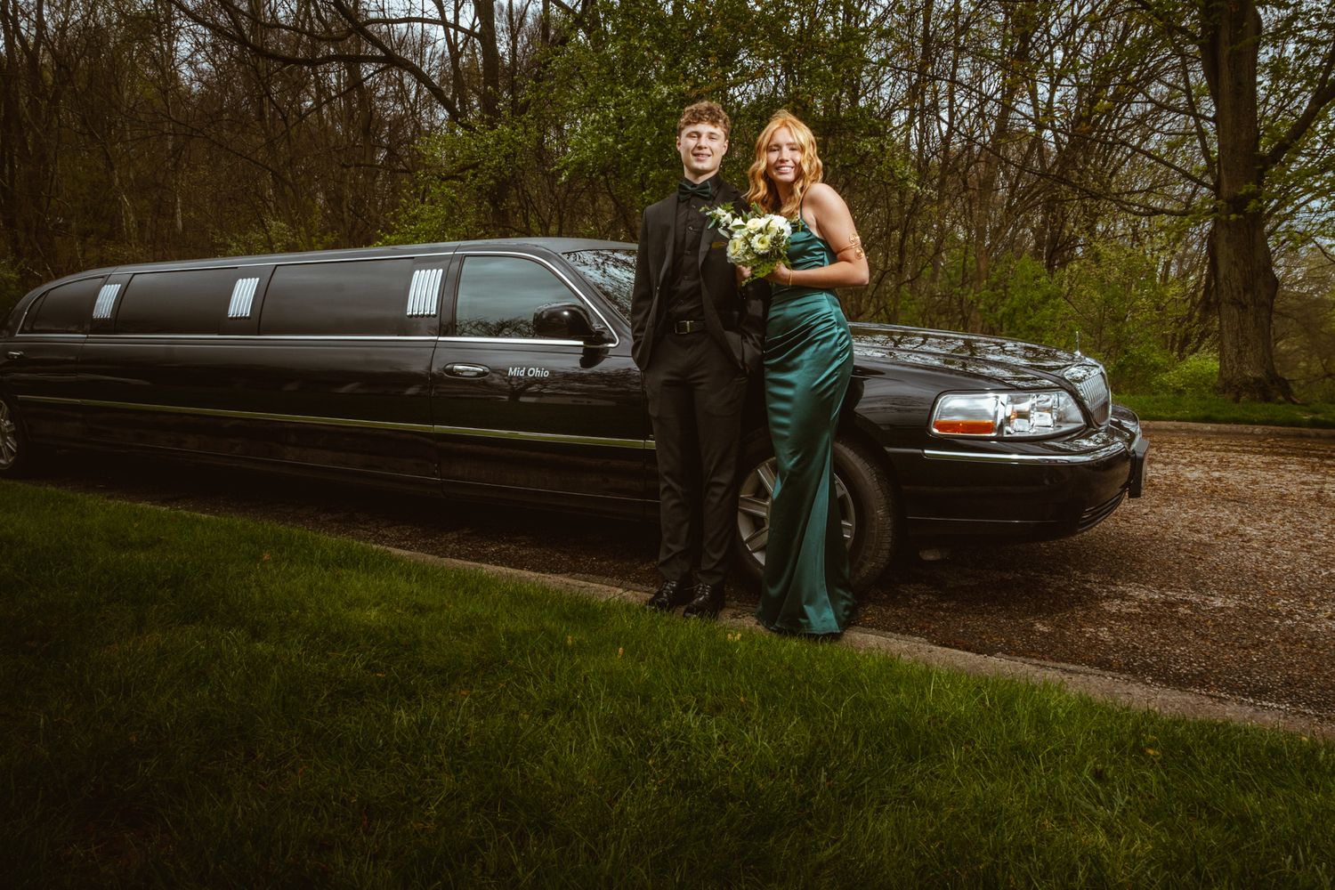 Couple poses by a black stretch limousine while holding flowers.