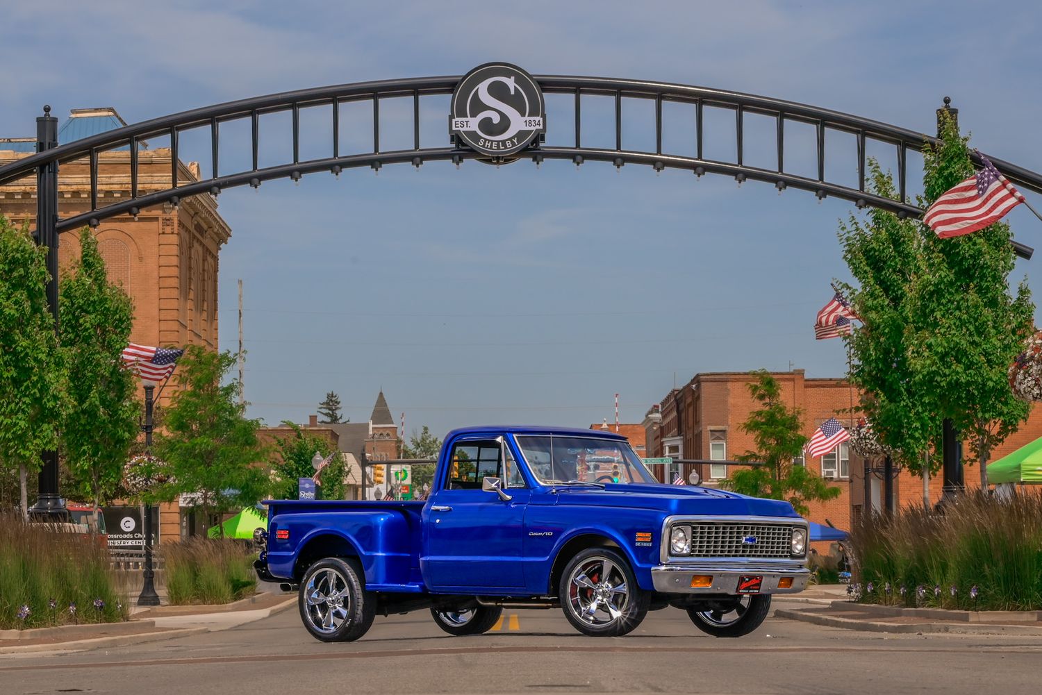 Custom blue Chevrolet C10 pickup truck with chrome wheels and lowered suspension driving through a brick-lined street.