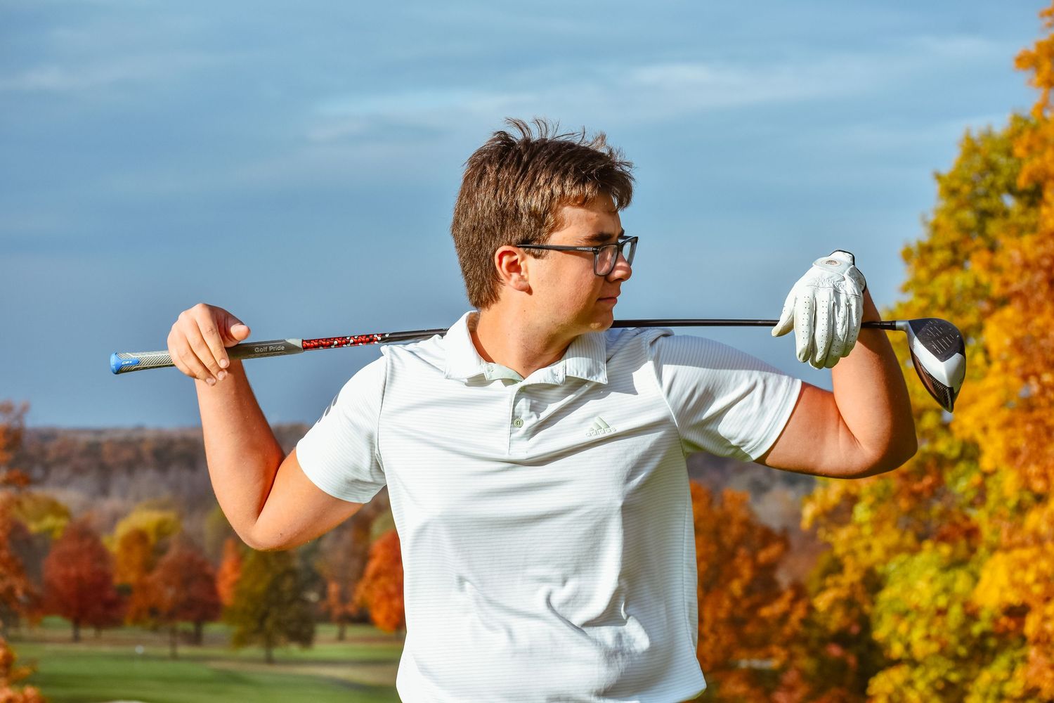 Golfer in white polo shirt stretches with club across shoulders against autumn foliage and blue sky background.
