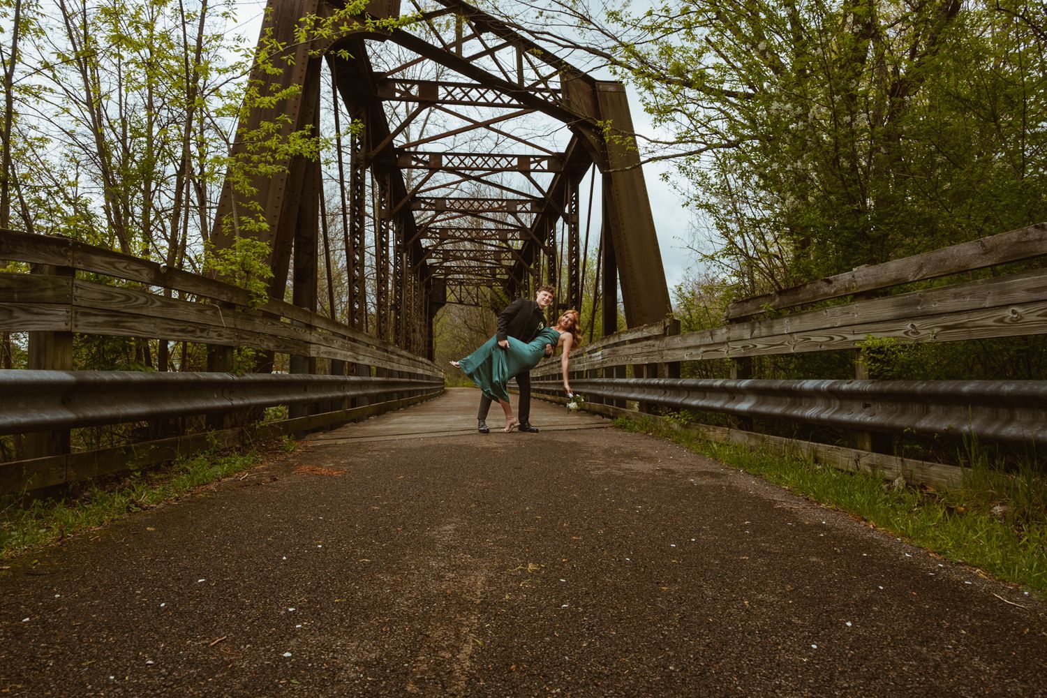 A figure stands on a rustic iron truss bridge surrounded by spring foliage on a dirt path.
