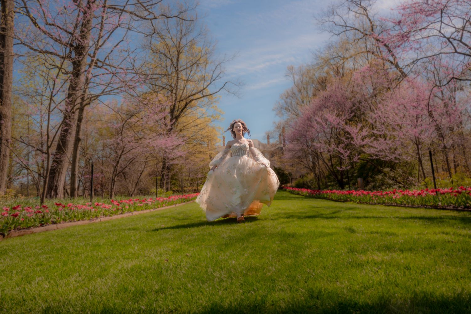 Bride in white gown twirling joyfully in a lush green park surrounded by blooming spring trees and tulips.