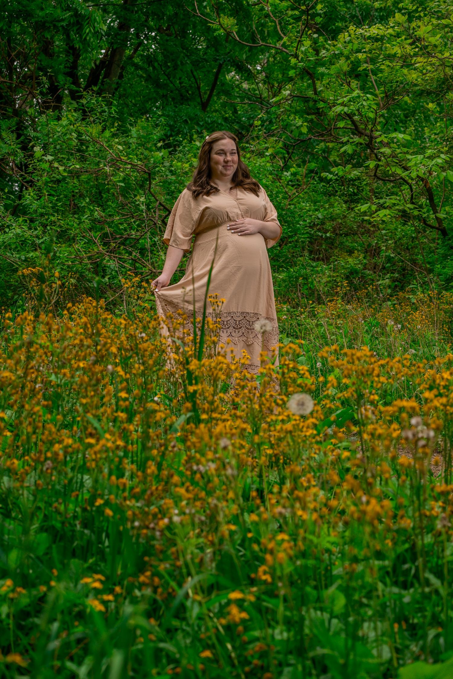 Pregnant woman in beige lace dress standing in wildflower meadow surrounded by lush green trees.