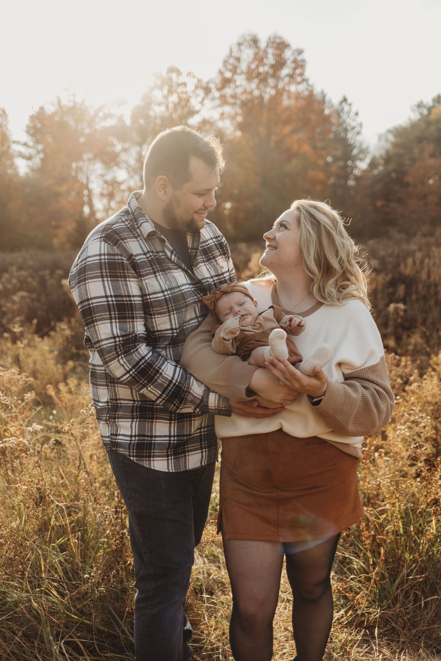 A couple laughs with their newborn in a golden field at sunset.