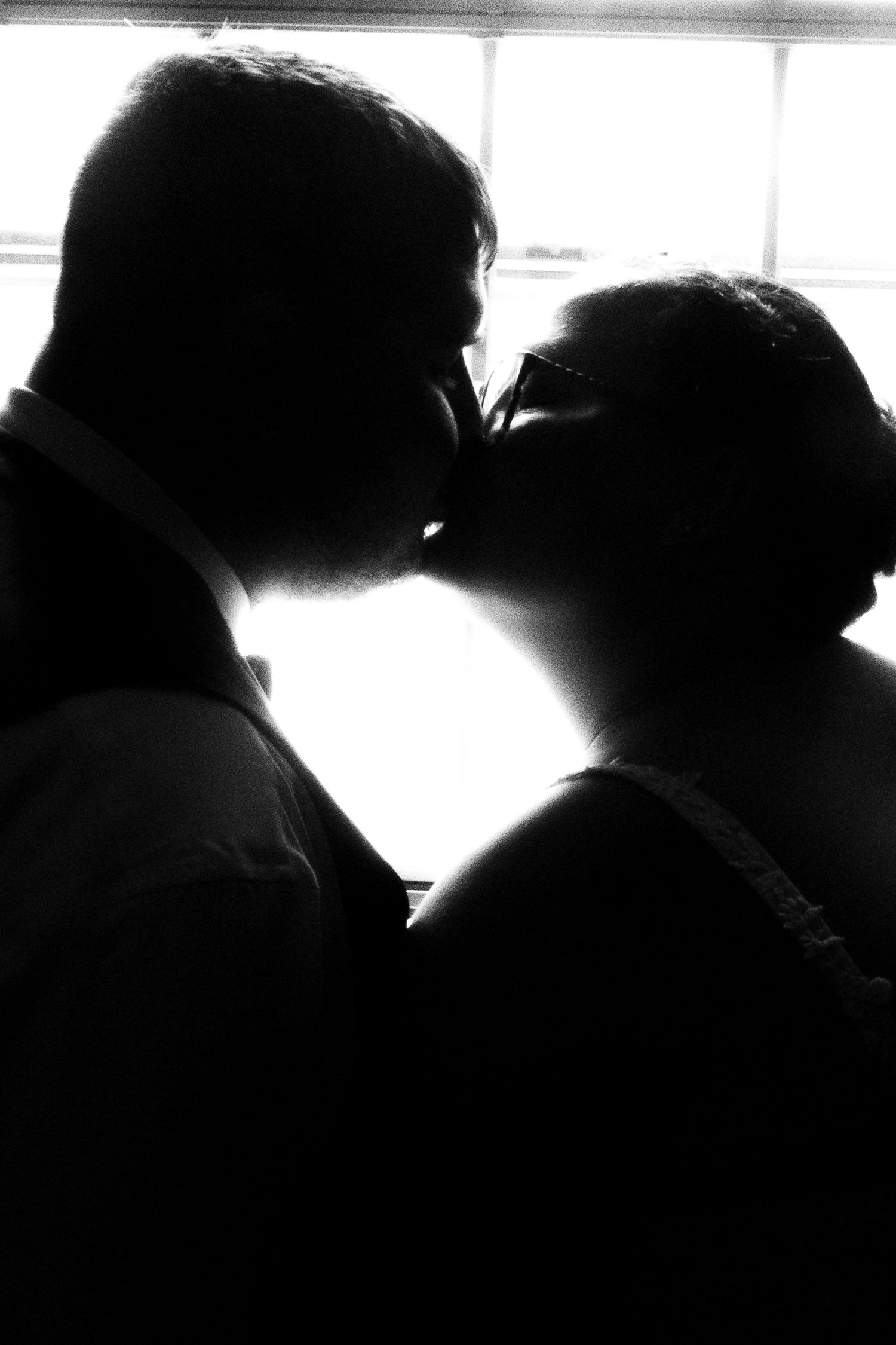Black and white silhouette of a couple sharing a romantic kiss backlit by a bright window.