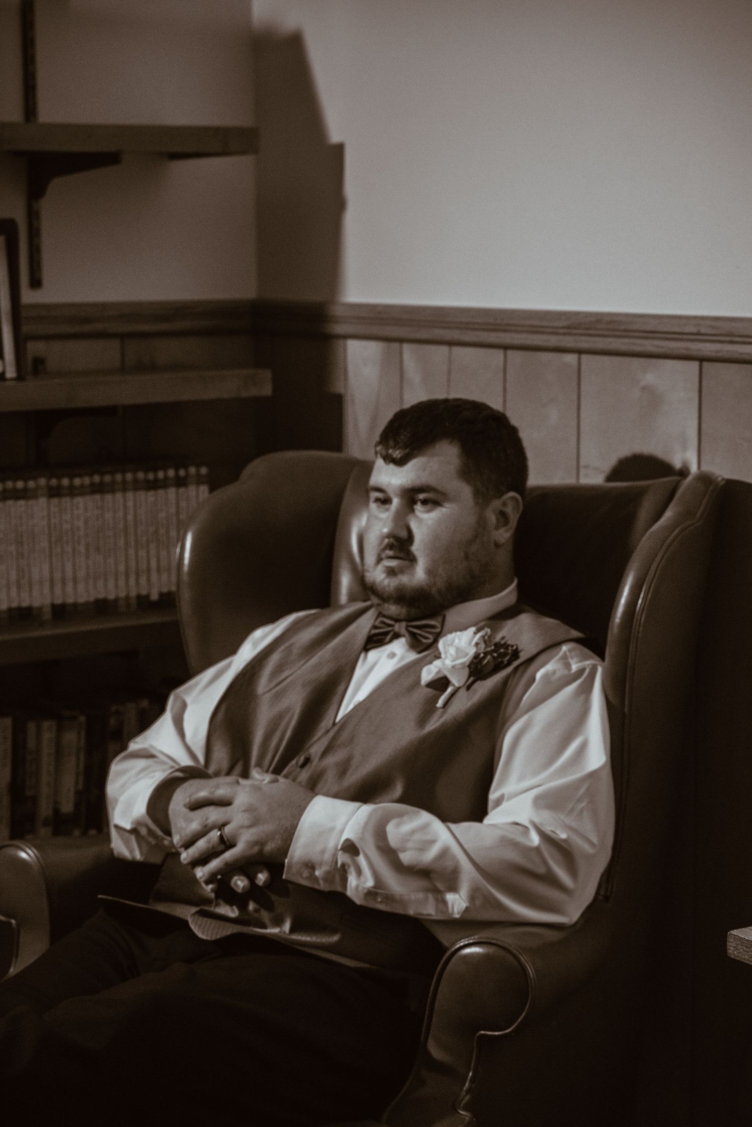 A groom in a vest and bow tie relaxes in a leather chair beside a bookshelf in a sepia-toned wedding photo.