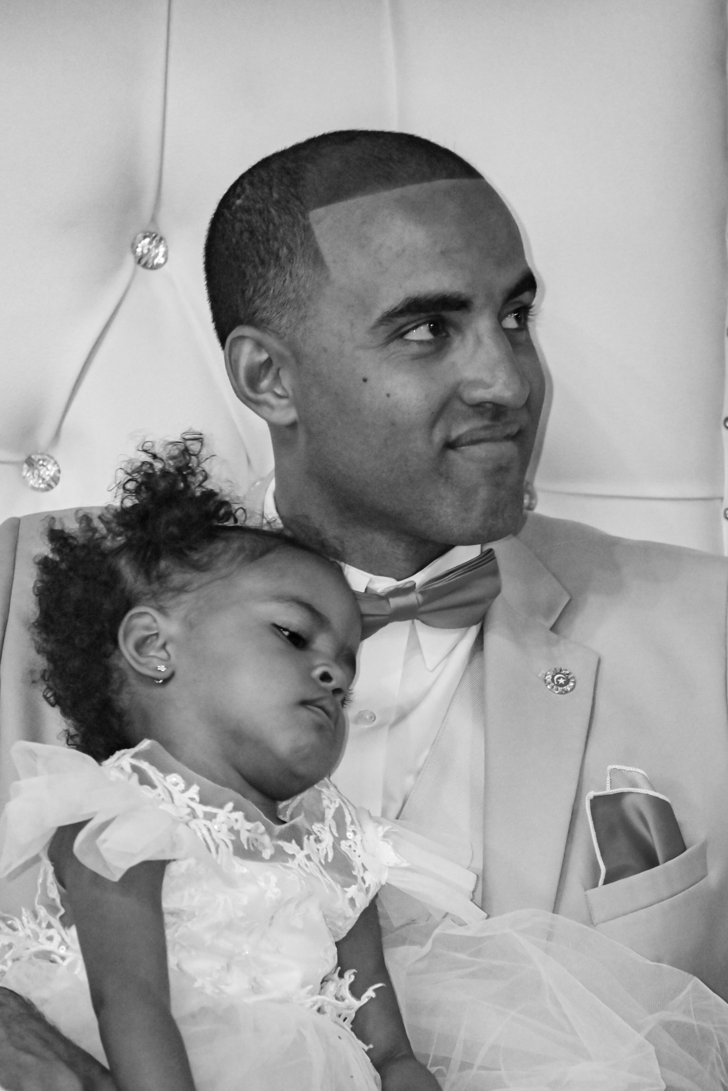 Black and white photo of a man in a bow tie suit holding a young girl in a white dress at a formal event.