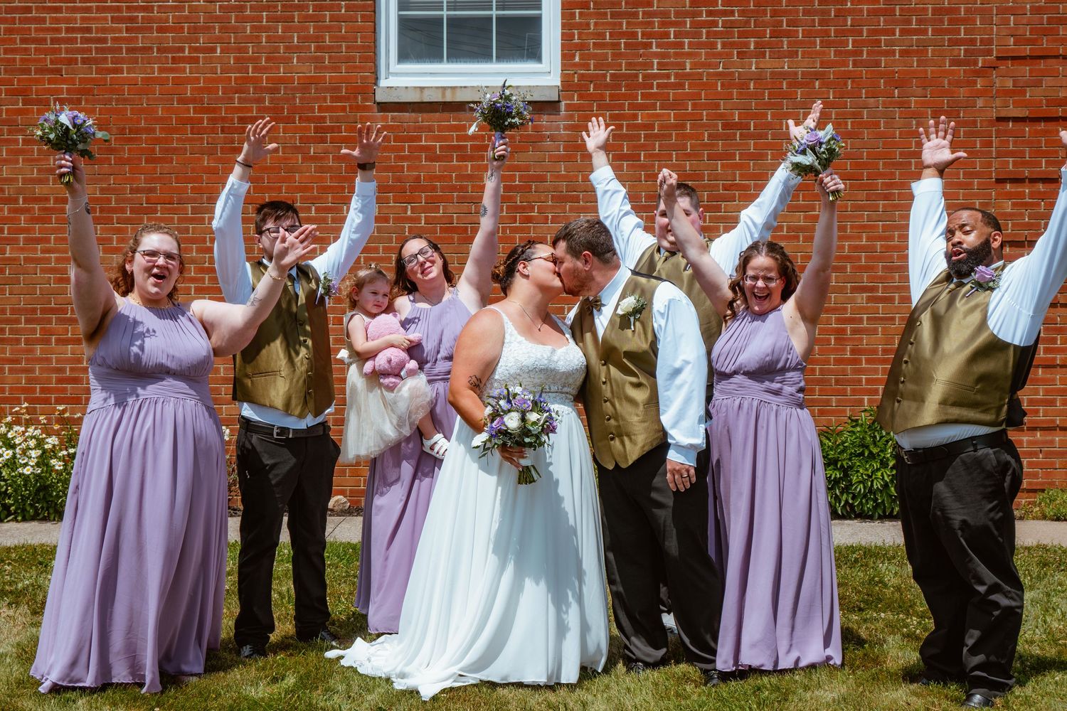 Wedding party celebrating outdoors, bridesmaids in lavender gowns, groomsmen in gold vests, couple kissing while guests cheer.