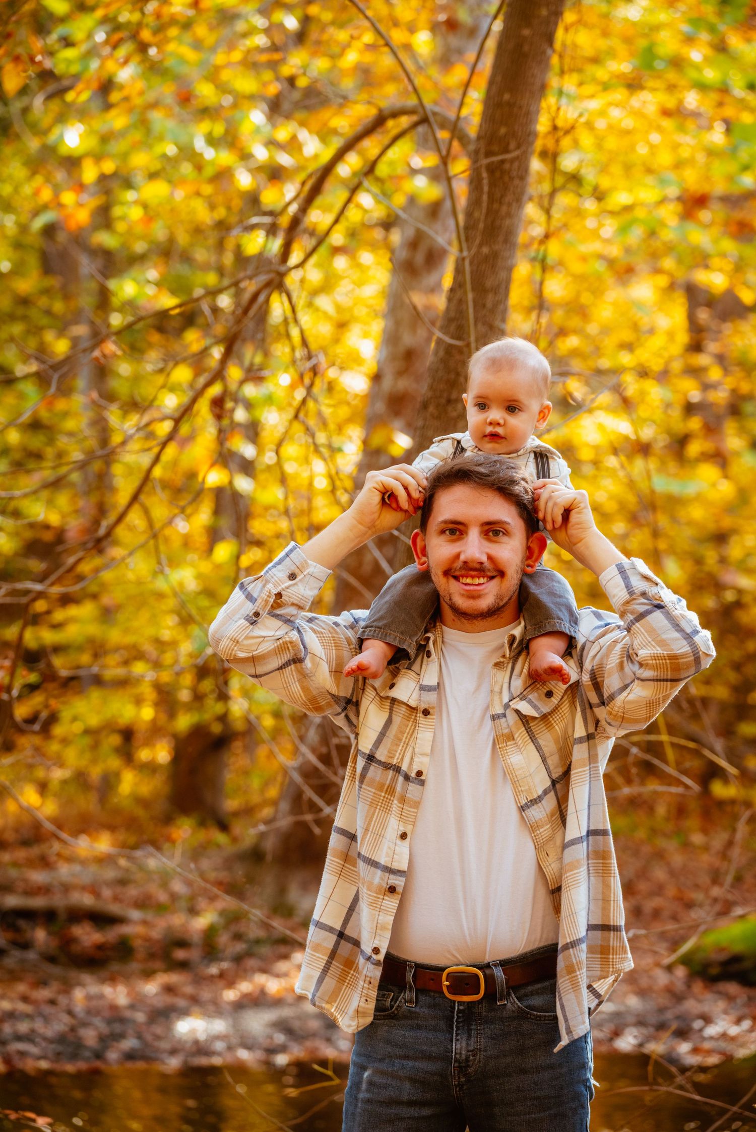 A smiling man carries a baby on his shoulders in an autumn forest with vibrant yellow foliage.