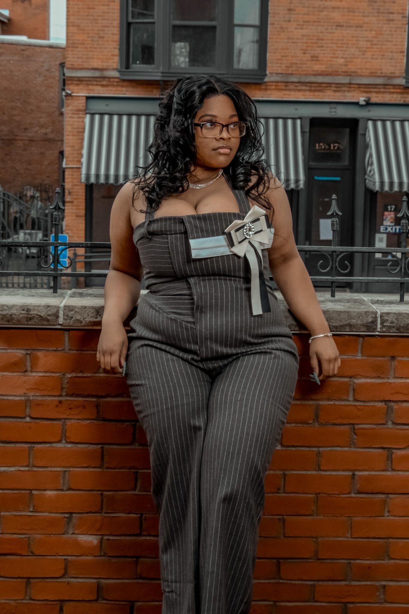 Woman in pinstripe jumpsuit with bow detail poses against brick wall in urban setting.