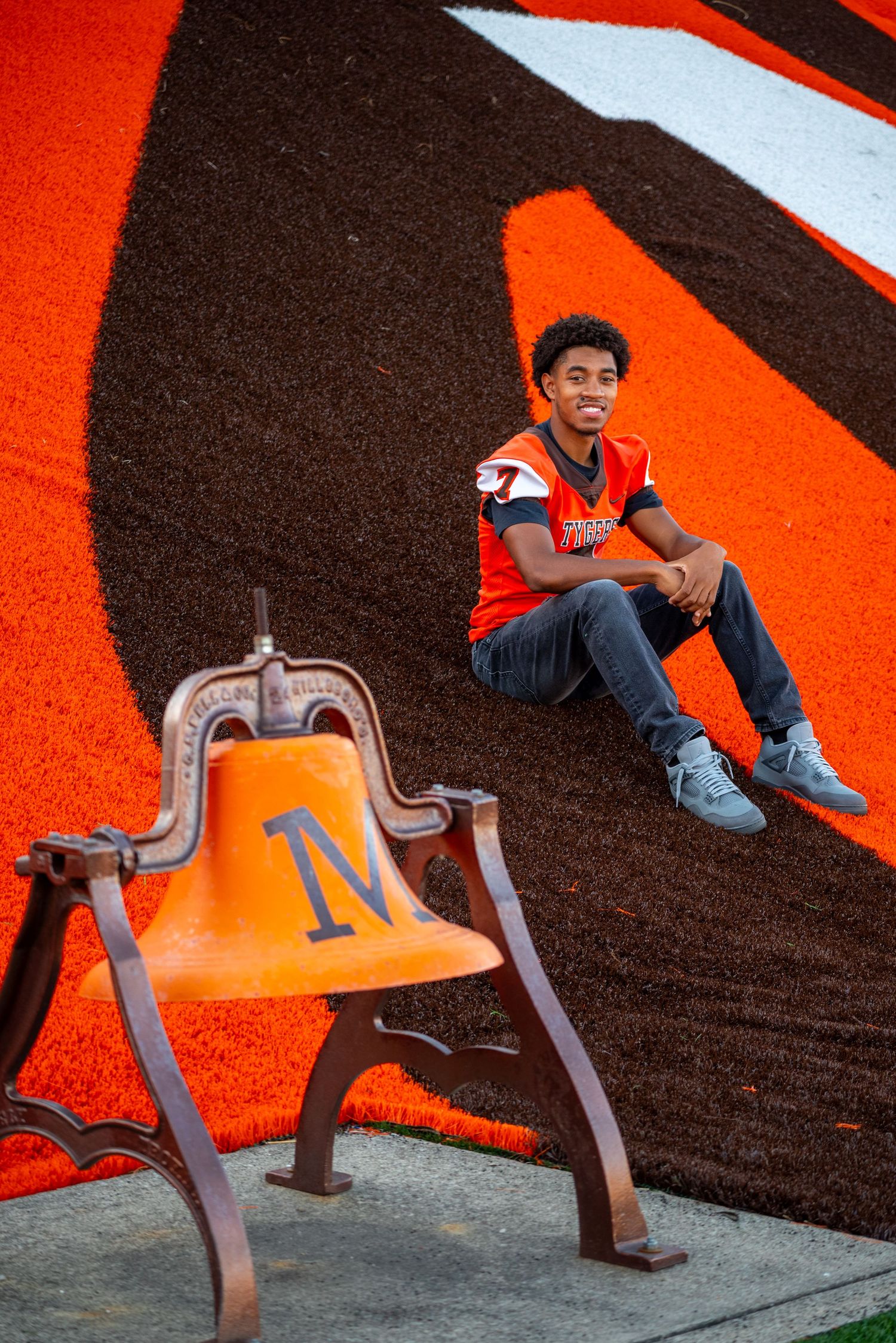 Young man in orange jersey sits on a vibrant orange and brown football field logo beside an orange bell with M initial.